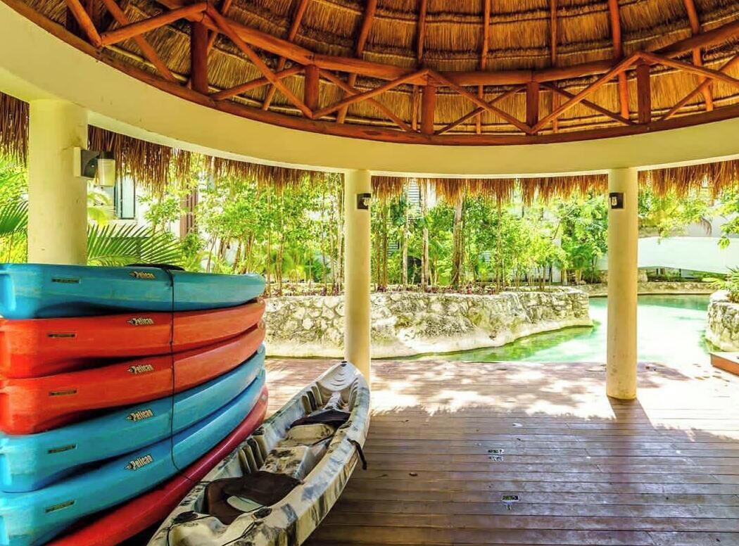 A covered outdoor area with a wooden deck, a thatched roof, and a view of a pool and greenery.