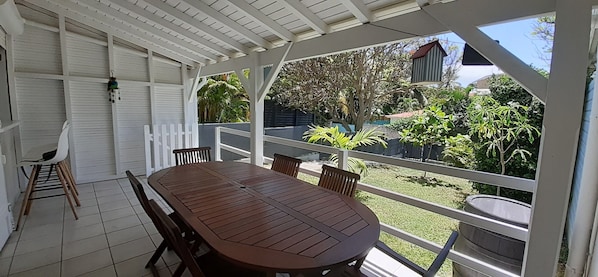 Une terrasse couverte avec une table et des chaises en bois, une clôture blanche et une vue sur un jardin où poussent différentes plantes, avec une maison en arrière-plan.