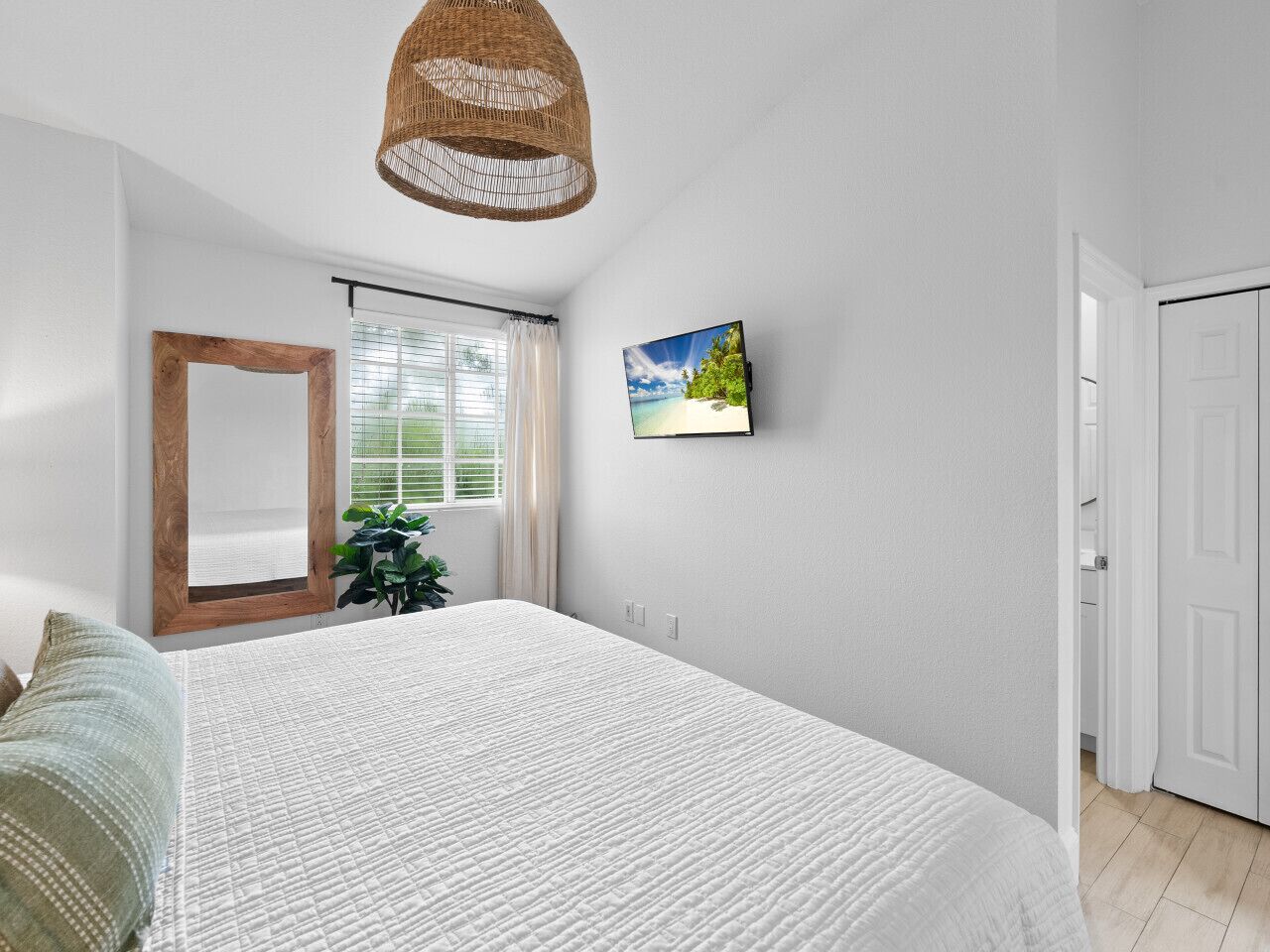 A bedroom with a large bed, a wooden-framed mirror, a window with blinds, a hanging wicker light fixture, and a wall-mounted picture.
