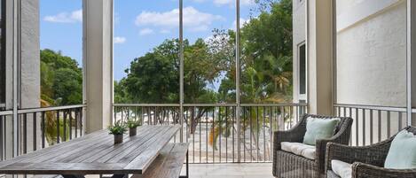 A covered patio with a wooden table and wicker furniture, overlooking a beach and palm trees.