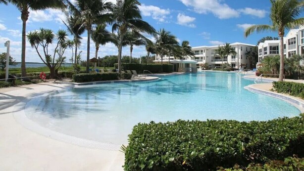 En pool vid ett semesterhotell, med palmer, en klarblå himmel och en byggnad i bakgrunden.