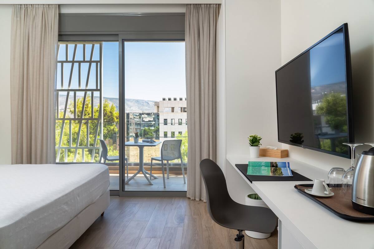 Een hotelkamer met een bed, een bureau met een computer, een televisie en een balkon met uitzicht op gebouwen en bomen.