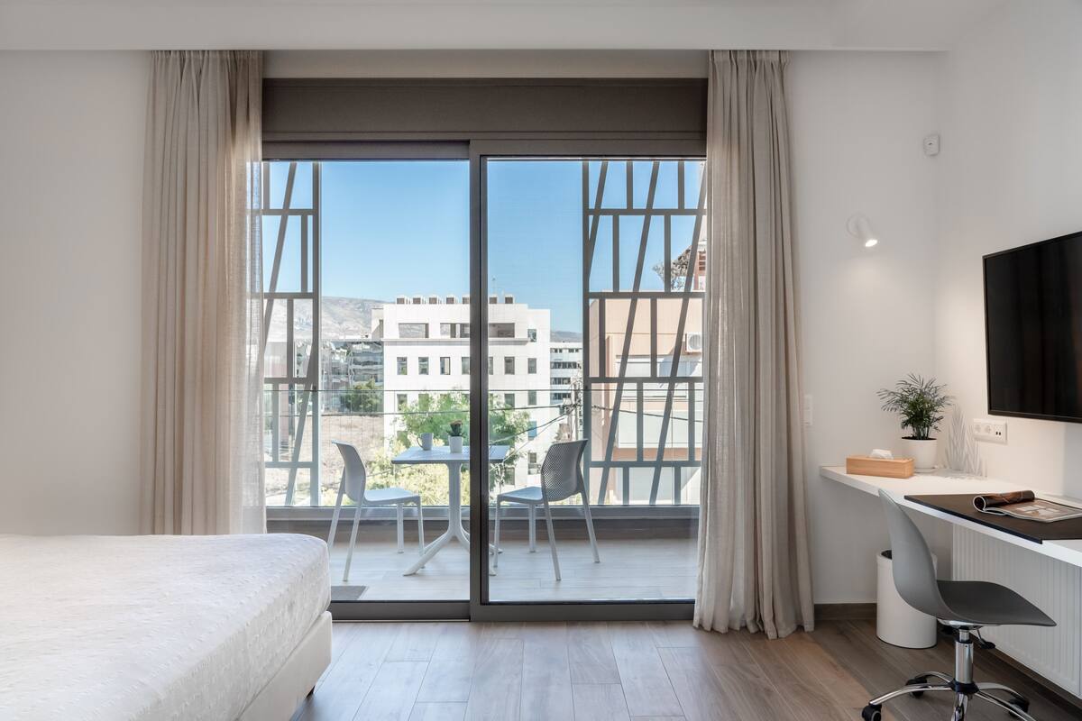 Een moderne hotelkamer met een grote schuifglazen deur naar een balkon, een bed, een bureau met stoel, en een aan de muur bevestigde televisie.