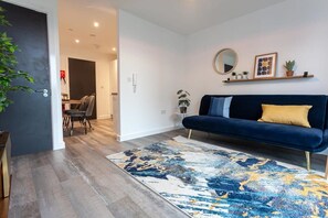 A modern living room with a dark blue sofa, a patterned rug, and a minimalist decor including a mirror and framed pictures on the wall.