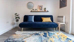 Ein modernes Wohnzimmer mit einem dunkelblauen Sofa, einem runden Spiegel, einem kleinen Beistelltisch mit Topfpflanze und einem dekorativen Regal mit Kleinigkeiten.