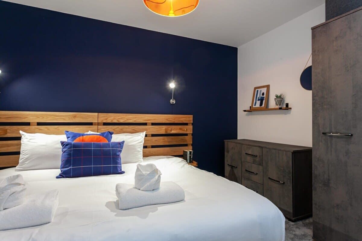 A modern hotel room with a wooden headboard, a large bed with white linens, a blue and orange pillow, and a bedside table with a lamp.