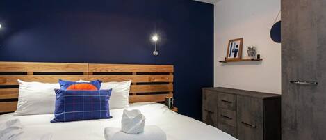 A modern hotel room with a wooden headboard, a large bed with white linens, a blue and orange pillow, and a bedside table with a lamp.