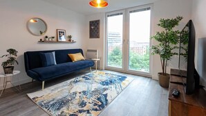 Ein modernes Wohnzimmer mit einem blauen Sofa, einem großen abstrakten Teppich, einem Fernseher und einem Balkon mit Blick auf Grünflächen.