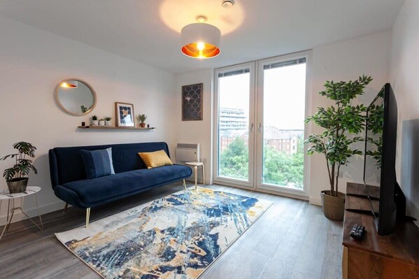 A modern living room with a blue sofa, a large abstract rug, a television, and a balcony with a view of greenery.