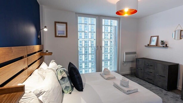 Ein modernes Schlafzimmer mit einem großen Bett, einem Holz-Kopfteil, einem Nachttisch, einer Kommode und einem Fenster mit Jalousien.