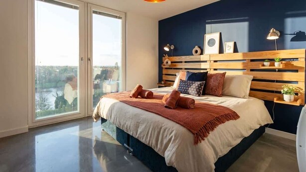 A modern bedroom with a large bed, a wooden headboard, a nightstand, and a view through a sliding door. - Bright Contemporary 2BED Apartment by Old Trafford (Manchester)