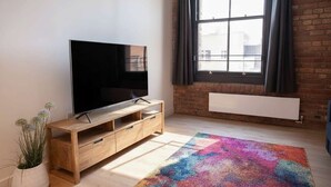 Een moderne woonkamer met een flatscreen televisie op een houten tv-meubel, een kleurrijk vloerkleed, een groot raam met gordijnen en een radiator.