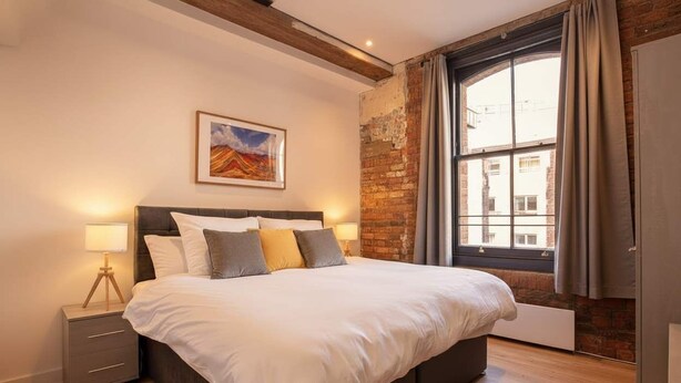 Een slaapkamer met een groot bed, een nachtkastje, een raam met gordijnen en een bakstenen muur.