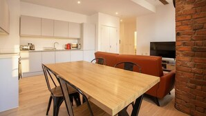 Een moderne keuken met witte kastjes, een houten eettafel met stoelen, een accentmuur van baksteen en een televisie in de woonkamer.