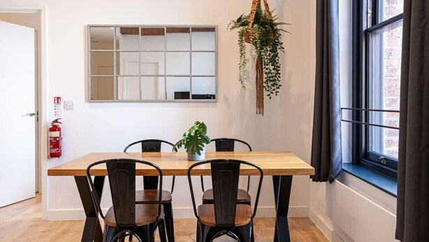 Een eetgedeelte met een houten tafel en zwarte stoelen, een hangplant, een raam met gordijnen en een brandblusser aan de muur.