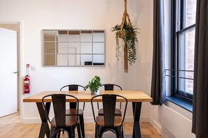 Ein Essbereich mit Holztisch und schwarzen Stühlen, einer hängenden Pflanze, einem Fenster mit Vorhängen und einem an der Wand befestigten Feuerlöscher.