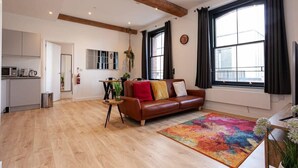 Een ruime woonkamer met een bruine leren bank, een kleurrijk vloerkleed en een groot raam met zwarte gordijnen.