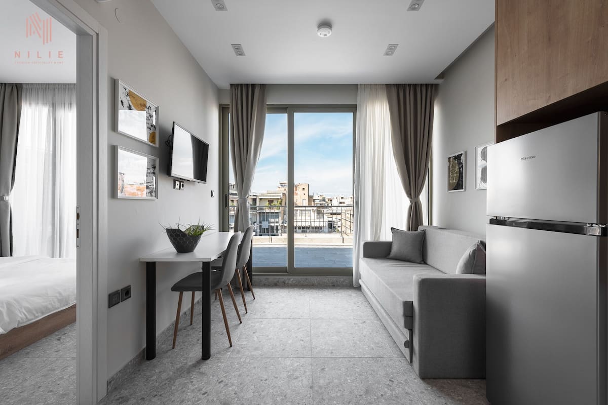 A modern, well-lit room with a dining area, a sofa, and a large window offering a view of the city.