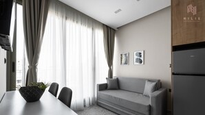 Ein modernes Hotelzimmer mit grauem Sofa, einem Esstisch mit Stühlen, einem schwarzen Kühlschrank und einem großen Fenster mit durchsichtigen Vorhängen.