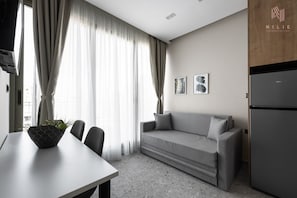 A modern, minimalist living room with a grey sofa, a dining table with chairs, and a black refrigerator. - Skysphere Nilie Hospitality MGMT (Thessaloniki)