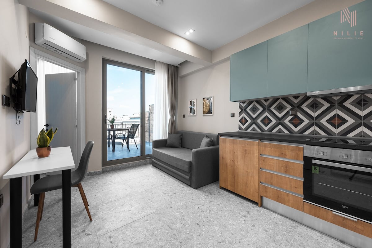 A modern kitchen with a built-in oven, a dining area with a table and chairs, a sofa, and a large sliding glass door leading to a balcony.