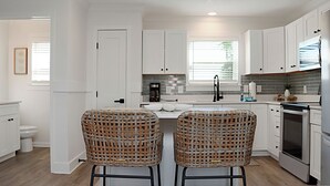 A modern kitchen with white cabinetry, a stainless steel oven, and two wicker bar stools.