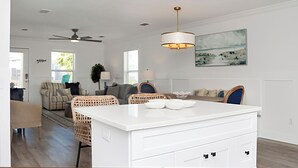 A modern living room with a white island, wicker chairs, a ceiling fan, and a painting on the wall.