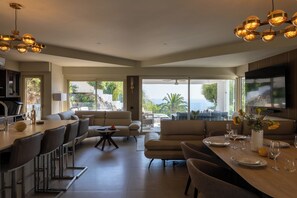 A modern living room with a dining area, large windows, and a view of the outdoors. - The Amazing Villa Boron! View of the ocean, pool, and hot tub (Nice)
