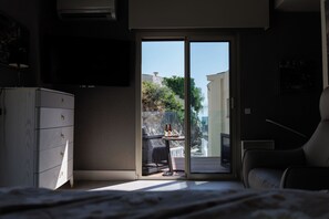 A hotel room with a bed, a desk, a chair, a minibar, and a sliding glass door leading to a balcony with a view of buildings and greenery. - The Amazing Villa Boron! View of the ocean, pool, and hot tub (Nice)