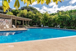 A swimming pool with lounge chairs, a covered area, and lush greenery in the background.