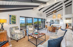 A spacious living area with a large window, a dining table, and a kitchen in the background.