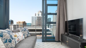 Un salon moderne avec un canapé gris, des coussins à motifs, une grande télévision et une vue sur la ville, visible à travers une porte vitrée.