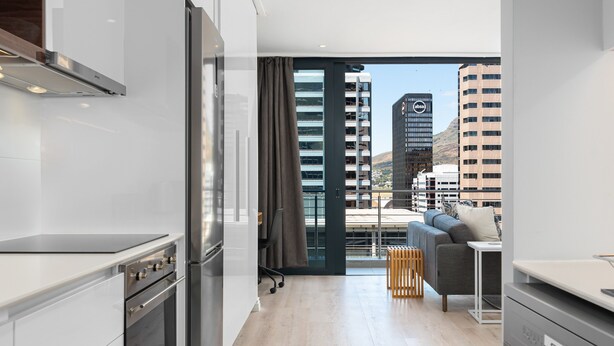 Une cuisine moderne équipée d’appareils électroménagers en acier inoxydable, un canapé gris et une vue sur les immeubles de la ville.