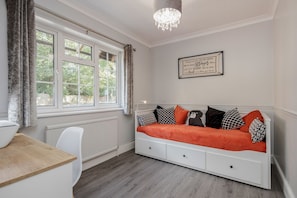 A room with a white sofa bed, a wooden table, a white chair, a window with curtains, and a chandelier.