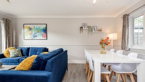 A modern living room with a blue sofa, white dining table, and chairs. There is a painting on the wall and a small shelf with decorative items. - Ascot Pet Friendly 4 Bed House Parking (Ascot)