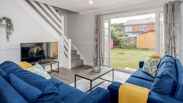 A modern living room with a blue sofa set, a flat-screen TV, a staircase, and a view of a garden through a sliding glass door. - Ascot Pet Friendly 4 Bed House Parking (Ascot)