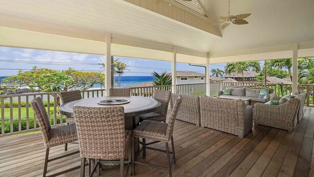 A covered outdoor deck with a round table and wicker furniture, overlooking a coastal view.