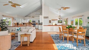 A spacious living area with a sofa, coffee table, and dining table with chairs. There are ceiling fans, a kitchen in the background, and large windows.