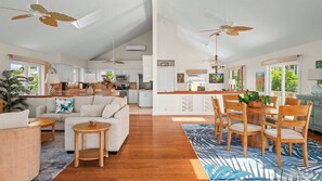 A spacious living area with a sofa, coffee table, and dining table with chairs. There are ceiling fans, a kitchen in the background, and large windows.