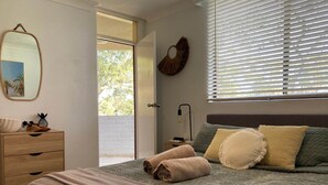 Une chambre à coucher avec un lit, une table de chevet, une lampe et une vue sur l’extérieur, visible à travers une porte ouverte.