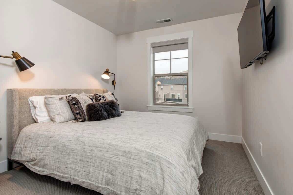 A bedroom with a bed, two bedside lamps, a window, and a TV mounted on the wall.
