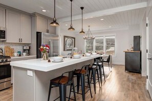 A modern kitchen with a central island, bar stools, pendant lighting, and a view through the window.