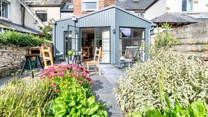 Una casa moderna con un patio que incluye una mesa y sillas, rodeada de exuberante follaje y plantas con flores.