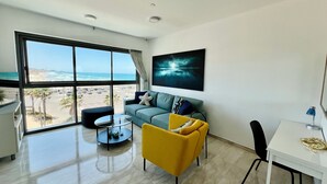 A modern living room with a large window overlooking a beach, a sofa, a yellow armchair, a small round table, and a desk with a lamp.