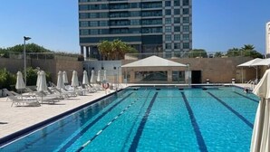 A swimming pool with clear blue water, surrounded by lounge chairs and umbrellas, with a tall building in the background.