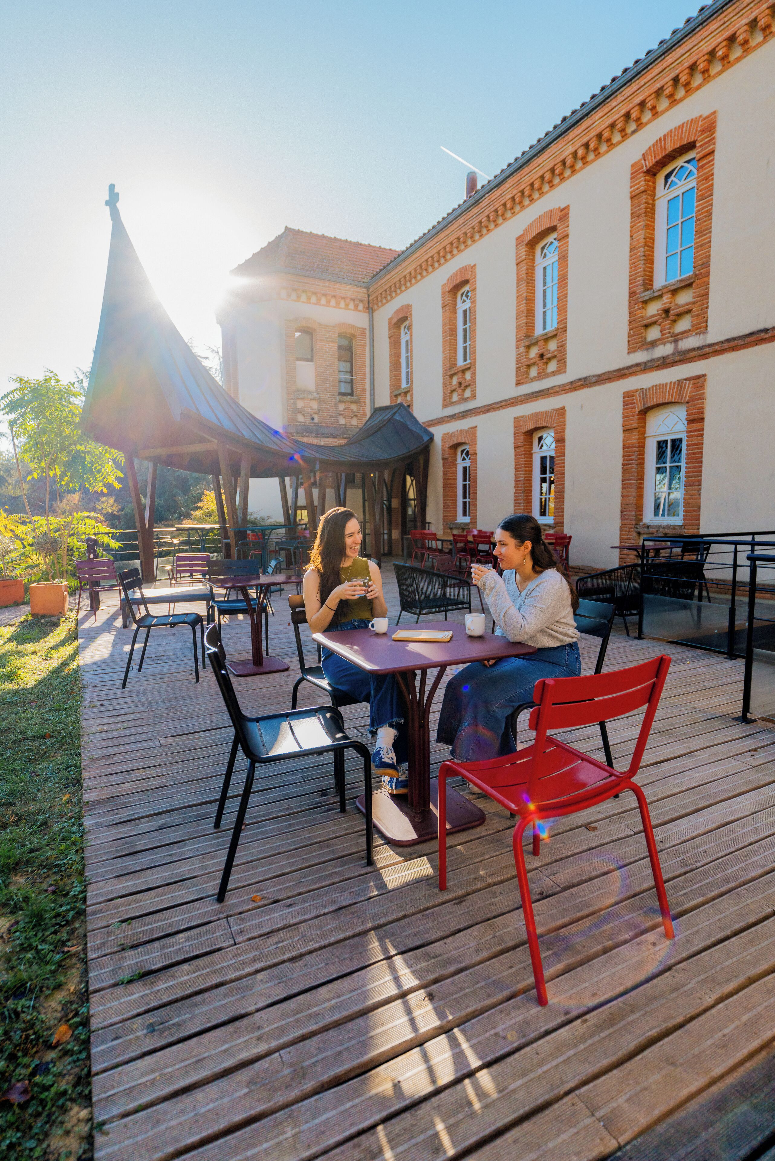 Un café en plein air avec des tables et des chaises en bois, un espace de repos couvert, et un bâtiment aux murs de briques et aux fenêtres qui se dessine à l’arrière-plan.