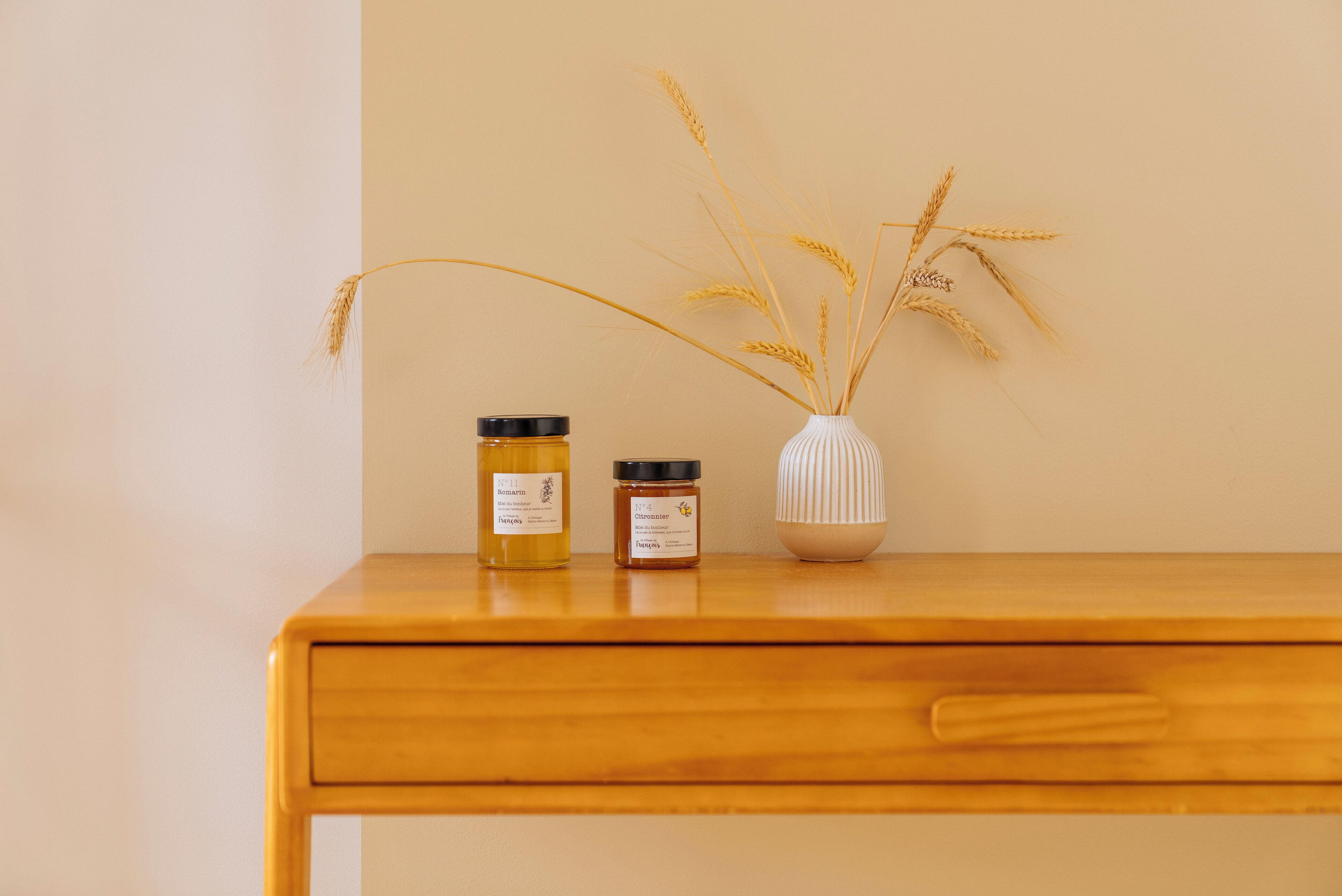A wooden table with two jars of honey and a vase with wheat stalks.