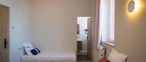 Un dormitorio pequeño y bien iluminado con dos camas, una ventana con persianas, una puerta y una lámpara.