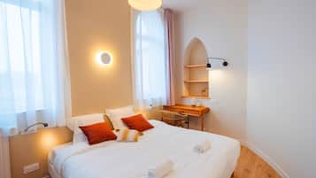 A hotel room with a large bed, a desk, and a chair. There is a window with sheer curtains and a wall-mounted shelf with a lamp.