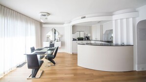 A modern kitchen with a curved island, a dining area with a glass table and chairs, and a staircase leading to an upper level.